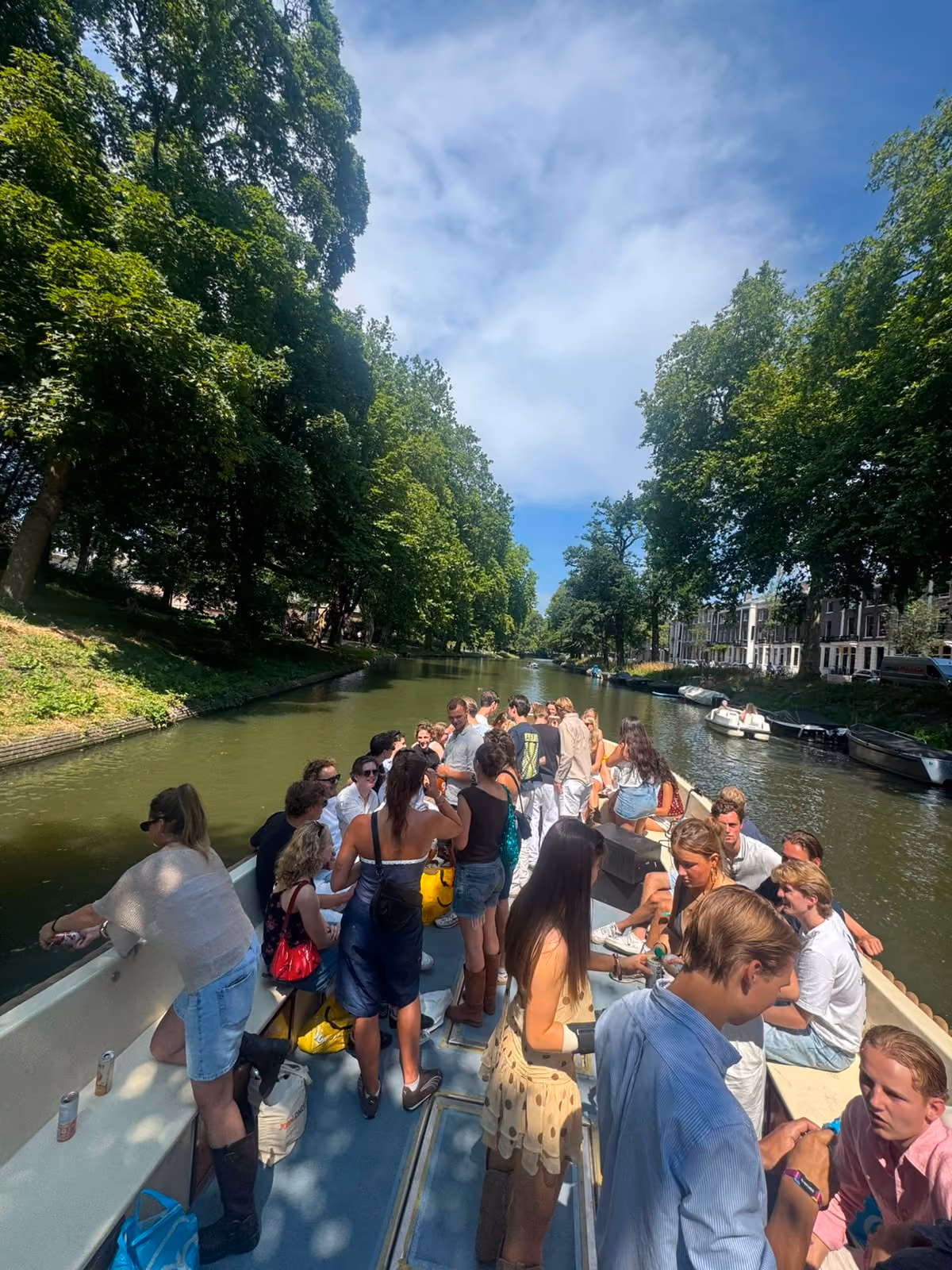 Large group of guests standing and chatting on an open boat on a tree-lined canal